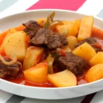 A bowl of beef stew with chunks of potatoes, carrots, and onions in a rich tomato broth, served on a striped pink and white background.