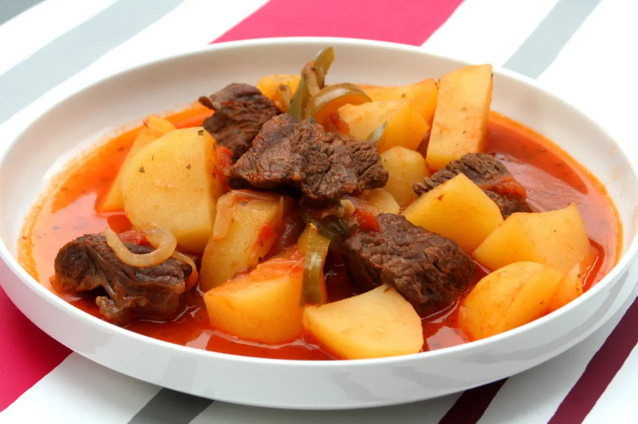 A bowl of beef stew with chunks of potatoes, carrots, and onions in a rich tomato broth, served on a striped pink and white background.