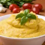 A bowl of creamy palenta garnished with a basil leaf, with tomatoes, garlic, and greens in the background