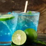 Two glasses of Blue Motorcycle Drink with lime wedges and ice cubes on a wooden table. The soda is a vibrant blue color and the lime wedges add a pop of green. The ice cubes are clinking against the sides of the glasses. The wooden table is a warm brown color and provides a contrast to the bright colors of the drinks.