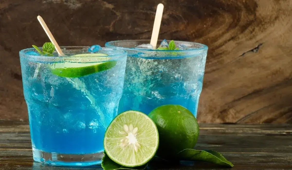 Two glasses of Blue Motorcycle Drink with lime wedges and ice cubes on a wooden table. The soda is a vibrant blue color and the lime wedges add a pop of green. The ice cubes are clinking against the sides of the glasses. The wooden table is a warm brown color and provides a contrast to the bright colors of the drinks.