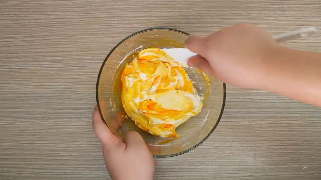 The image shows a hand using a spatula to mix Butter Slime in a clear glass bowl.