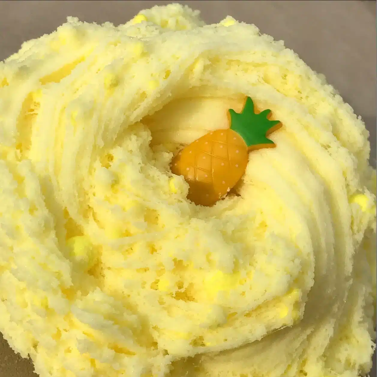 The image showcases a close-up of a swirled yellow confection, possibly a cloud dough slime, with a small, decorative pineapple-shaped piece nestled in the center.