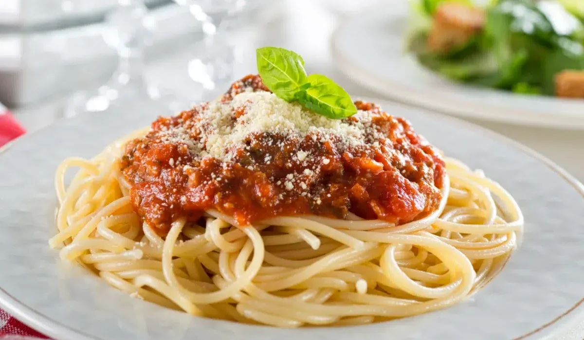 The image depicts a plate of Spaghetti In 10 Steps topped with a generous portion of marinara sauce, garnished with a fresh basil leaf and a sprinkle of grated Parmesan cheese.