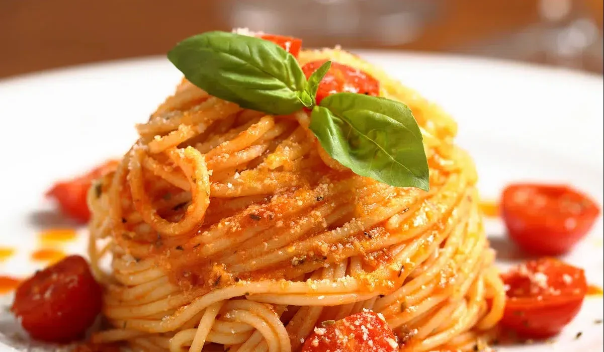 The image shows a beautifully presented plate of simple spaghetti pasta. The pasta is twirled into a high nest shape and topped with a bright green basil leaf, which adds a fresh accent.