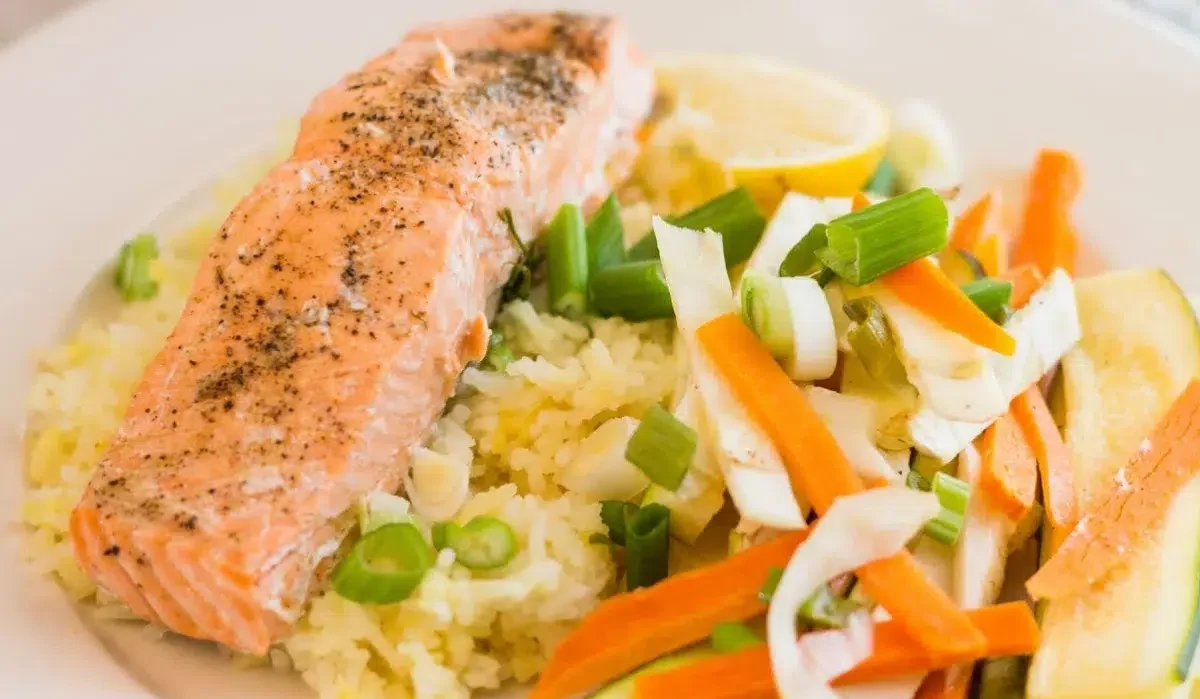 The image shows a plate of food consisting of a grilled salmon fillet seasoned with pepper, served alongside a bed of yellow rice and a variety of lightly cooked vegetables made by Wyse Guide.