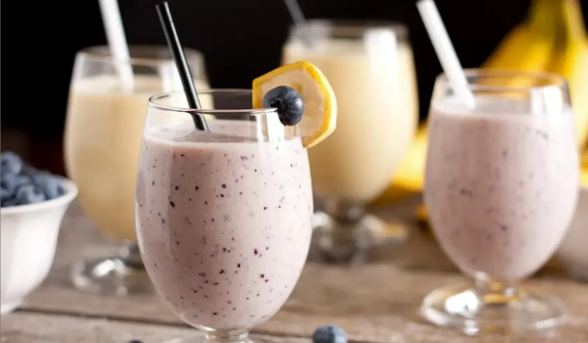 The image features three glasses of a creamy, Herbalife shake with one glass in the foreground adorned with a slice of lemon and a single blueberry on the rim.