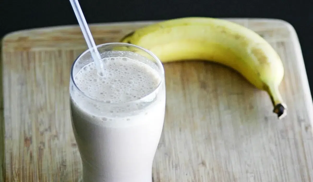 The image shows a tall glass of a creamy, frothy Herbalife shake with a straw, placed on a wooden cutting board.