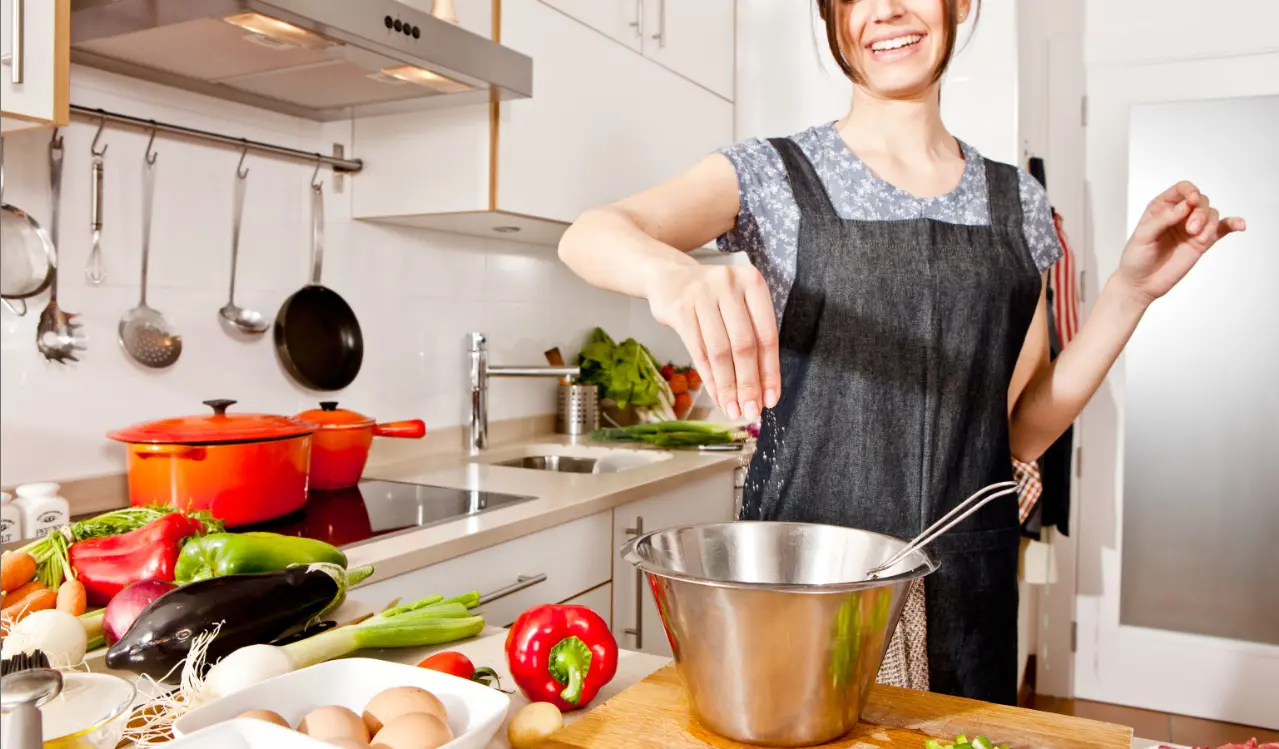 The image captures a cheerful woman in a kitchen, sprinkling an ingredient into a stainless steel mixing bowl for Publix apron