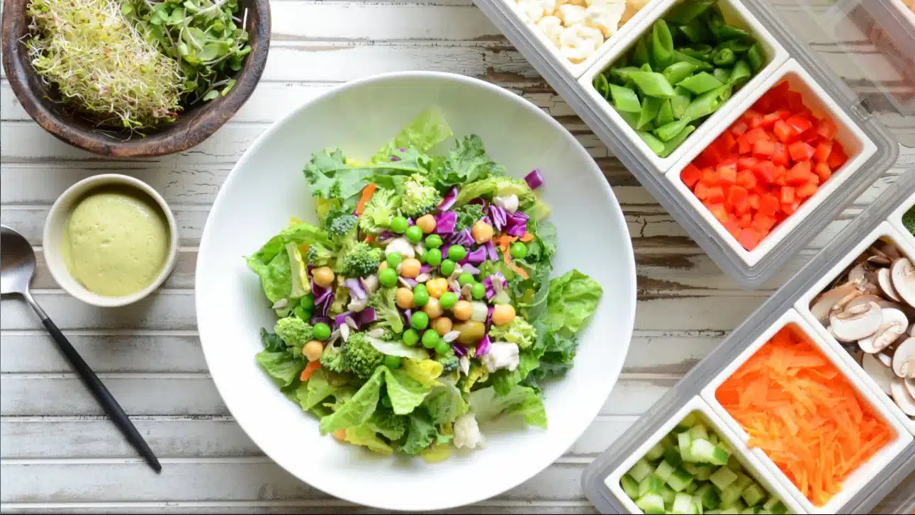 The image is from ninja speedi recipes features a fresh, colorful salad in a white bowl, which includes a variety of vegetables such as green lettuce, broccoli, purple cabbage, carrots, peas, and chickpeas. Next to the bowl is a small dish of creamy green dressing and a pair of black chopsticks. On the right, there are containers with organized, chopped vegetables ready to be added to a salad or used for meal preparation, including snow peas, red bell peppers, mushrooms, carrots, and cucumbers. The setting suggests a focus on healthy eating and meal preparation, with a wooden background that adds a rustic touch to the presentation.