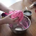 The image shows a person's hands kneading a vibrant pink substance, which resembles homemade cloud slime without borax, over a metal bowl.