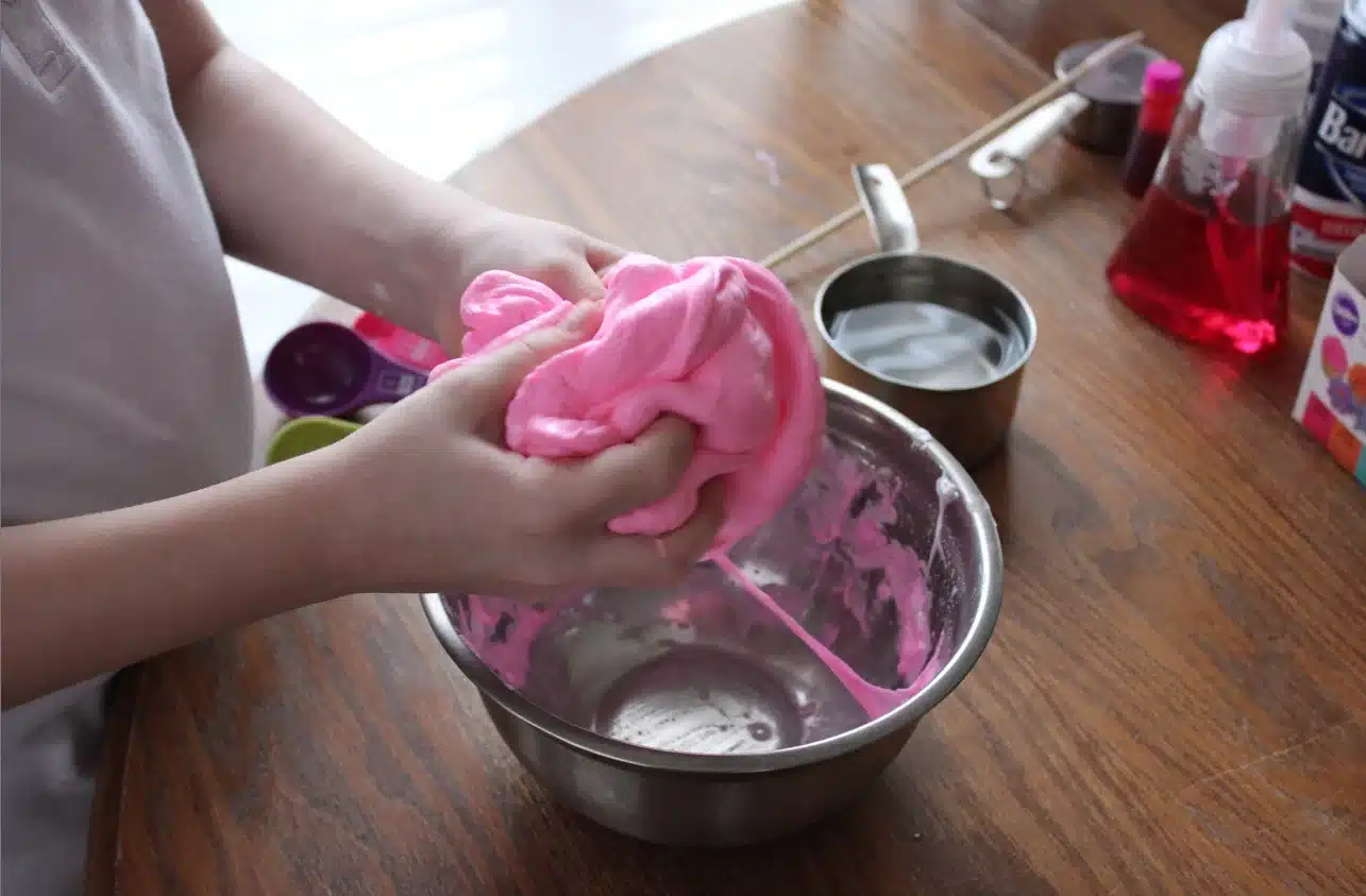The image shows a person's hands kneading a vibrant pink substance, which resembles homemade cloud slime without borax, over a metal bowl.