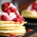 The image shows a close-up of a plate with a stack of Kylie Jenner Pancakes topped with whipped cream and a generous serving of strawberry compote.