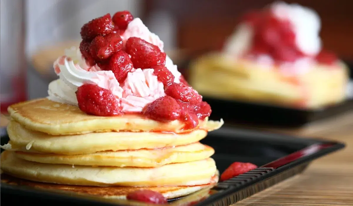 The image shows a close-up of a plate with a stack of Kylie Jenner Pancakes topped with whipped cream and a generous serving of strawberry compote.