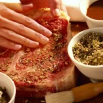 The image captures a close-up of hands seasoning a large raw Meat Church steak with a blend of spices.