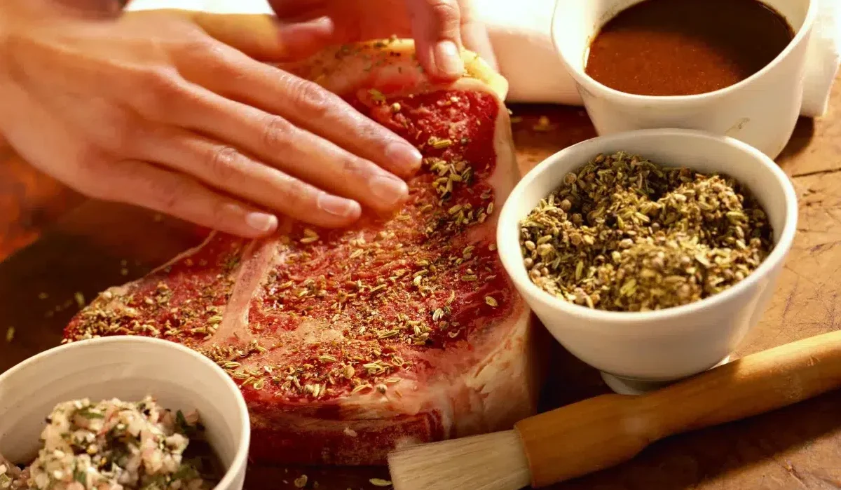 The image captures a close-up of hands seasoning a large raw Meat Church steak with a blend of spices.