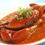 The image depicts a cooked crab in a reddish-crab barrack sauce, garnished with slices of red chili and green herbs, likely cilantro, presented on a white plate.