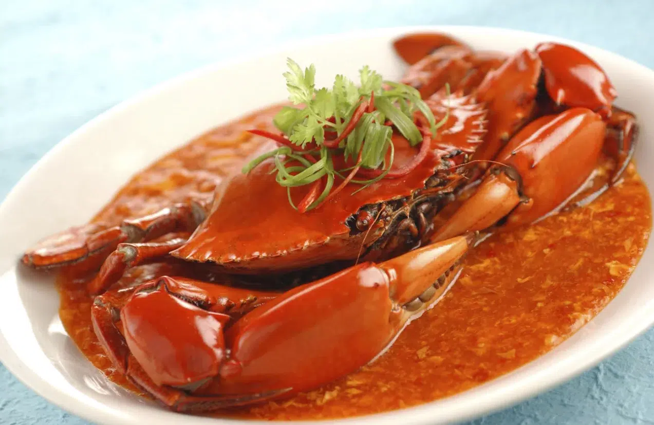 The image depicts a cooked crab in a reddish-crab barrack sauce, garnished with slices of red chili and green herbs, likely cilantro, presented on a white plate.
